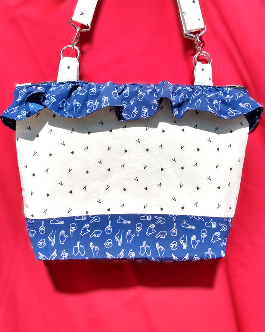 avant du sac signora. Le corps du sac est en lin blanc à motifs fanions triangulaires bleu marine. Le bas du sac est en coton bleu foncé à motifs mains blanches parlant en langue des signes. Une bande froncée du même tissu est présente sur le haut du sac. Les anses, même tissu que le corps du sac, sont accrochées via des anneaux et des mousquetons.